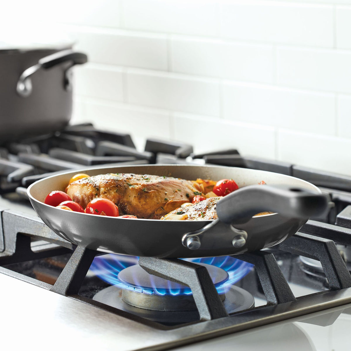 Chicken cooking in a frying pan on a gas stove with tomatoes.
