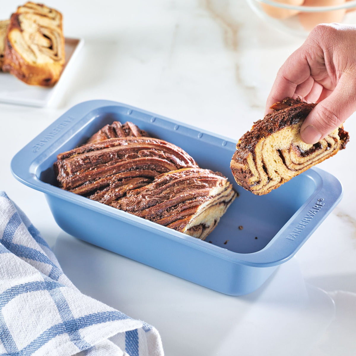 Person lifting a slice of braided chocolate bread from a blue baking dish.