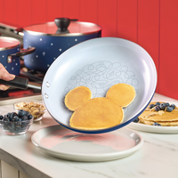 Pancake in a pan shaped like a mouse head, with berries and pans nearby.