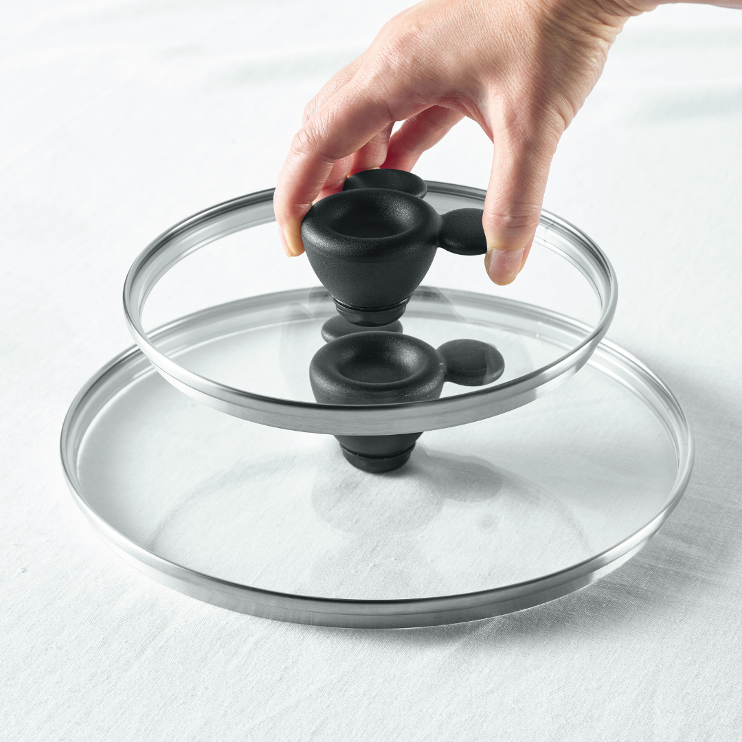 Hand lifting a glass pot lid with a black handle from a stack.