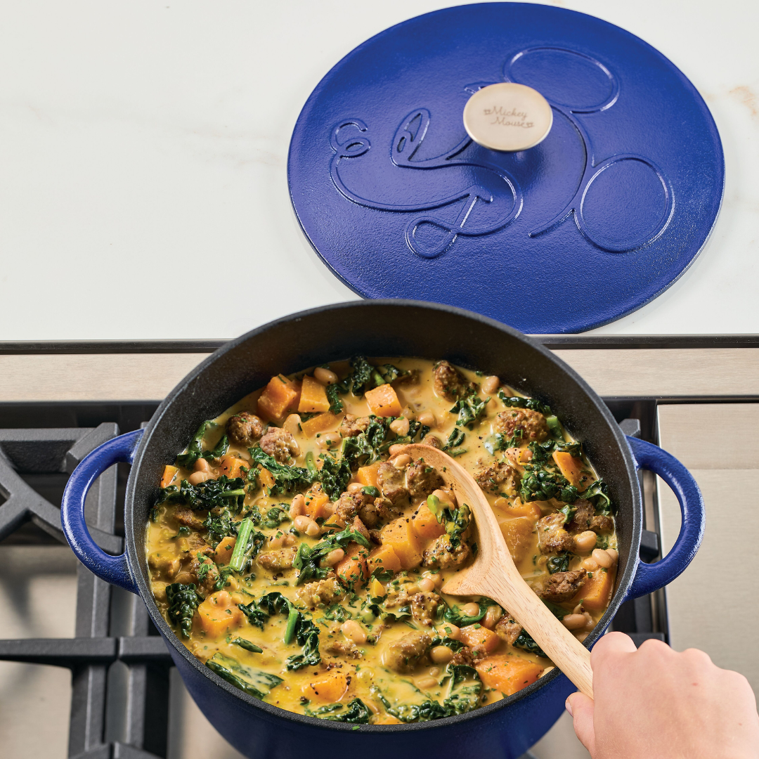 Pot of stew with vegetables and sausage on a stove, blue lid beside it.