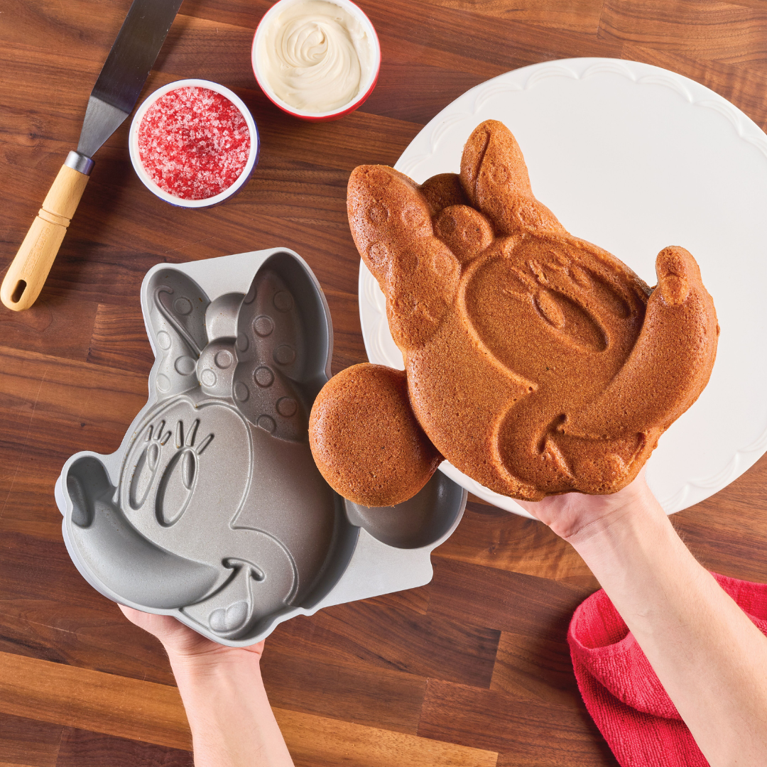 Hands holding a Disney character-shaped cake of Minnie Mouse next to its Minnie Mouse 3D Cake Pan on a wooden table.
