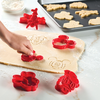A hand uses the Bake with Mickey Mouse 8-Piece Cookie Cutter and Stamp Set on dough, with more cookies resting on a baking sheet.