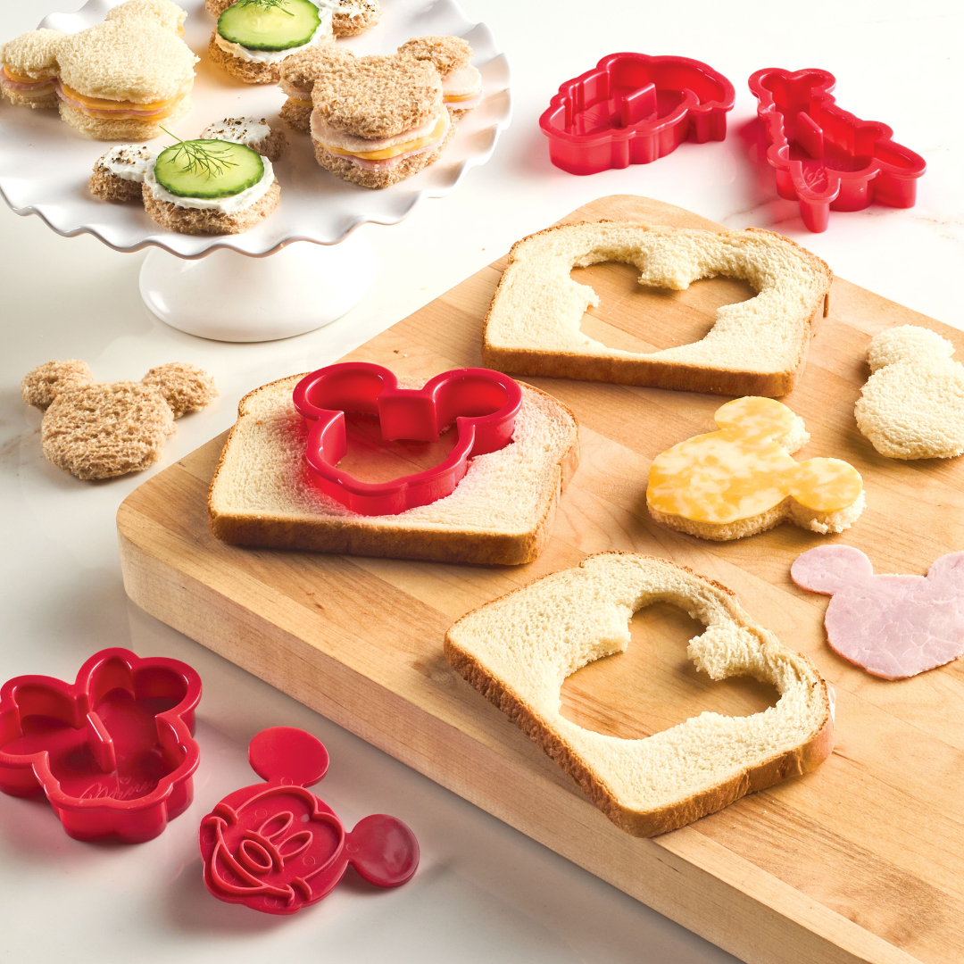 Slices of bread featuring mouse-shaped cutouts, crafted with a cutter from the Bake with Mickey Mouse 8-Piece Cookie Cutter and Stamp Set, are displayed on a wooden board next to vibrant red cutters.