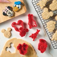 On the table, you'll find cookies and cutters from the Bake with Mickey Mouse 8-Piece Cookie Cutter and Stamp Set, showcasing the beloved Mickey and Friends. The baking set creates a charming atmosphere as dough is prepared to be shaped with these enchanting Disney cookie cutters, soon to be part of a collection of freshly baked treats that add a magical touch to your baking experience.