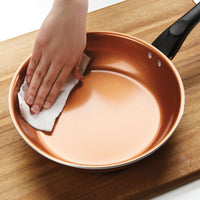 Hand wiping a copper frying pan with a paper towel on a wooden surface.
