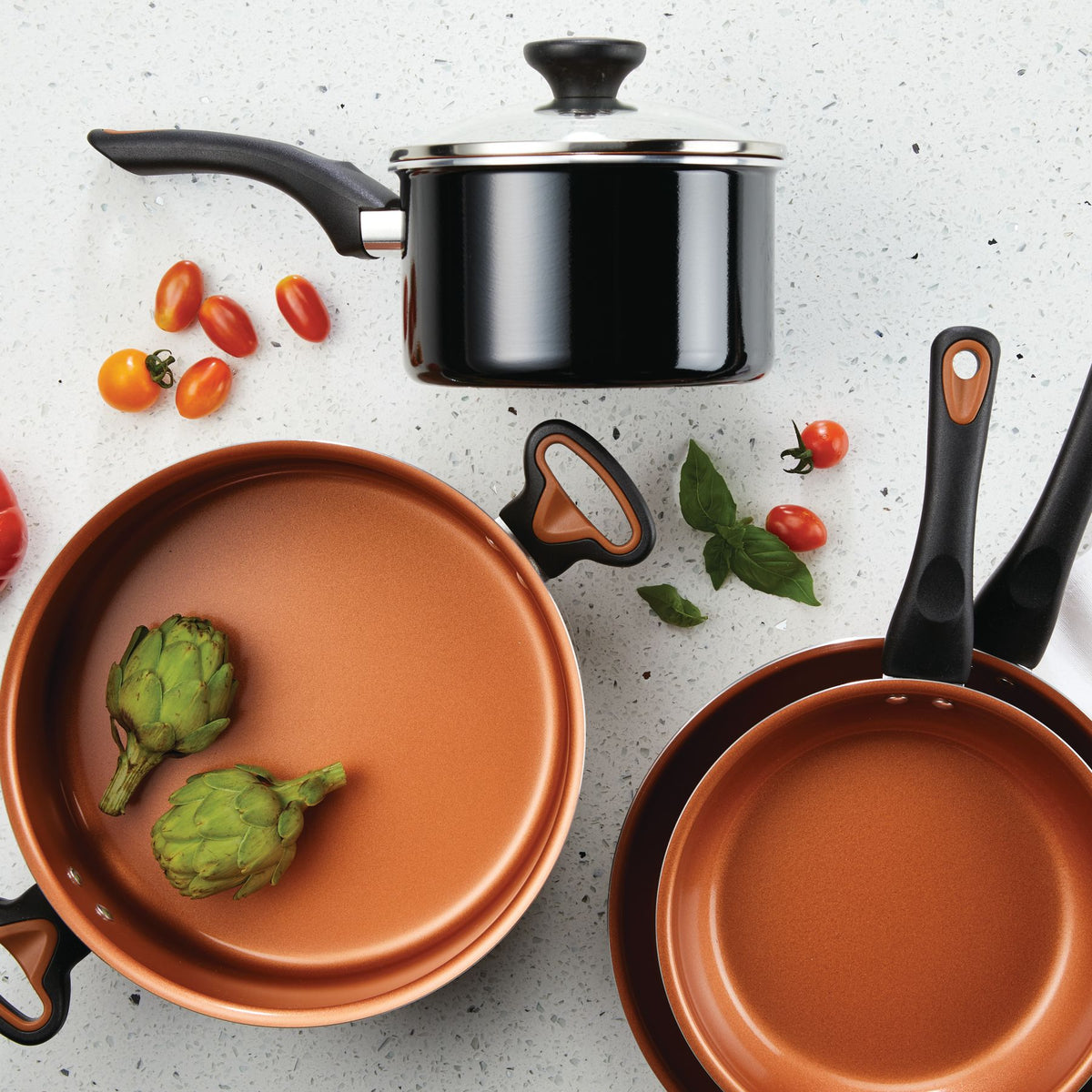 Copper cookware with artichokes and tomatoes on a white countertop.