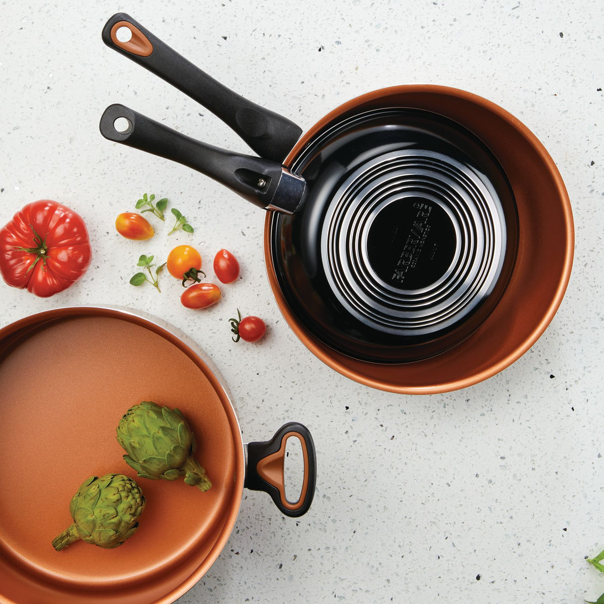 Two copper pans with tomatoes and artichokes on a granite countertop.