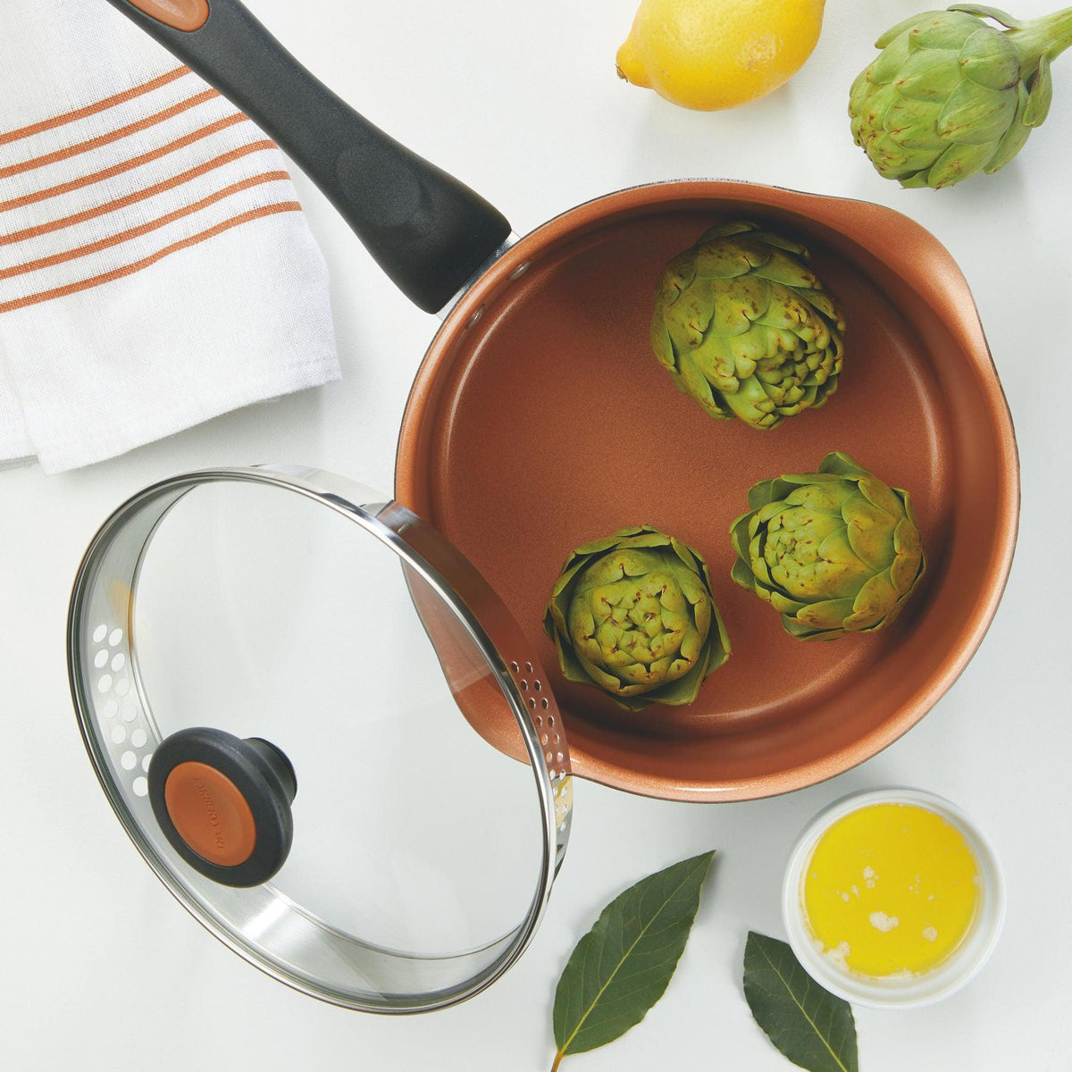 Copper pan with artichokes, a lemon, and butter on a white surface.