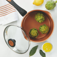 Cookware with artichokes, lemon, butter, and bay leaves on a white surface.