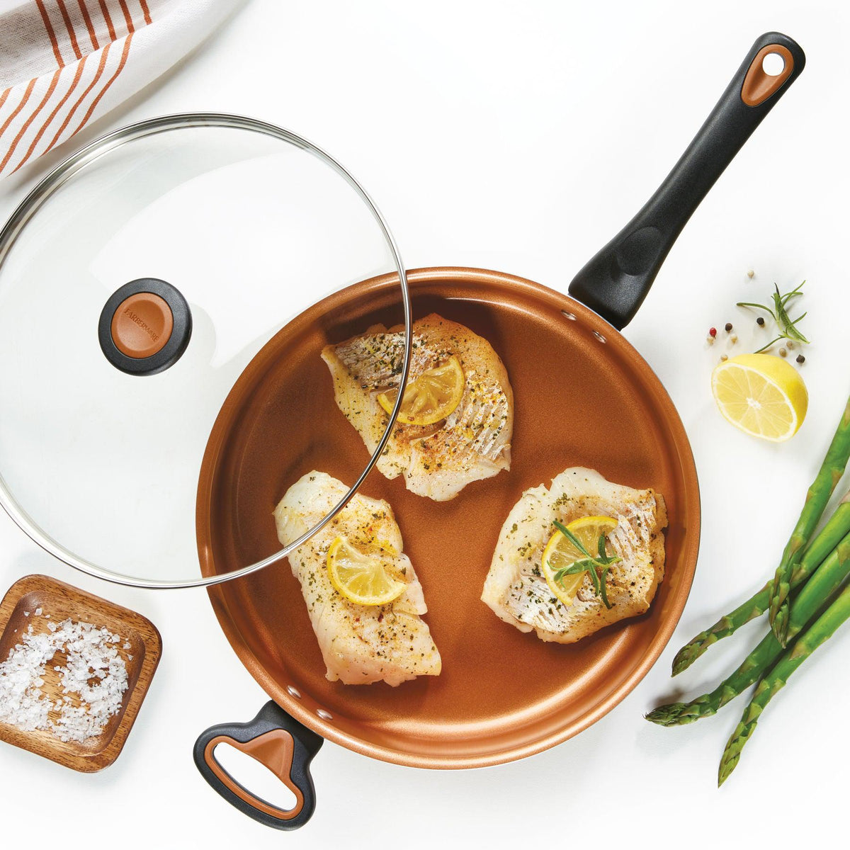 Fish fillets with lemon in a frying pan, asparagus, lemon, and salt nearby.