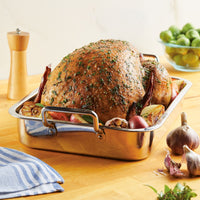 A roasted turkey sits in the 17" x 12.25" Stainless Steel Roaster with Rack, surrounded by garlic, onions, and herbs on a wooden table. This roaster features an oven-safe design and includes a removable rack for convenient serving.