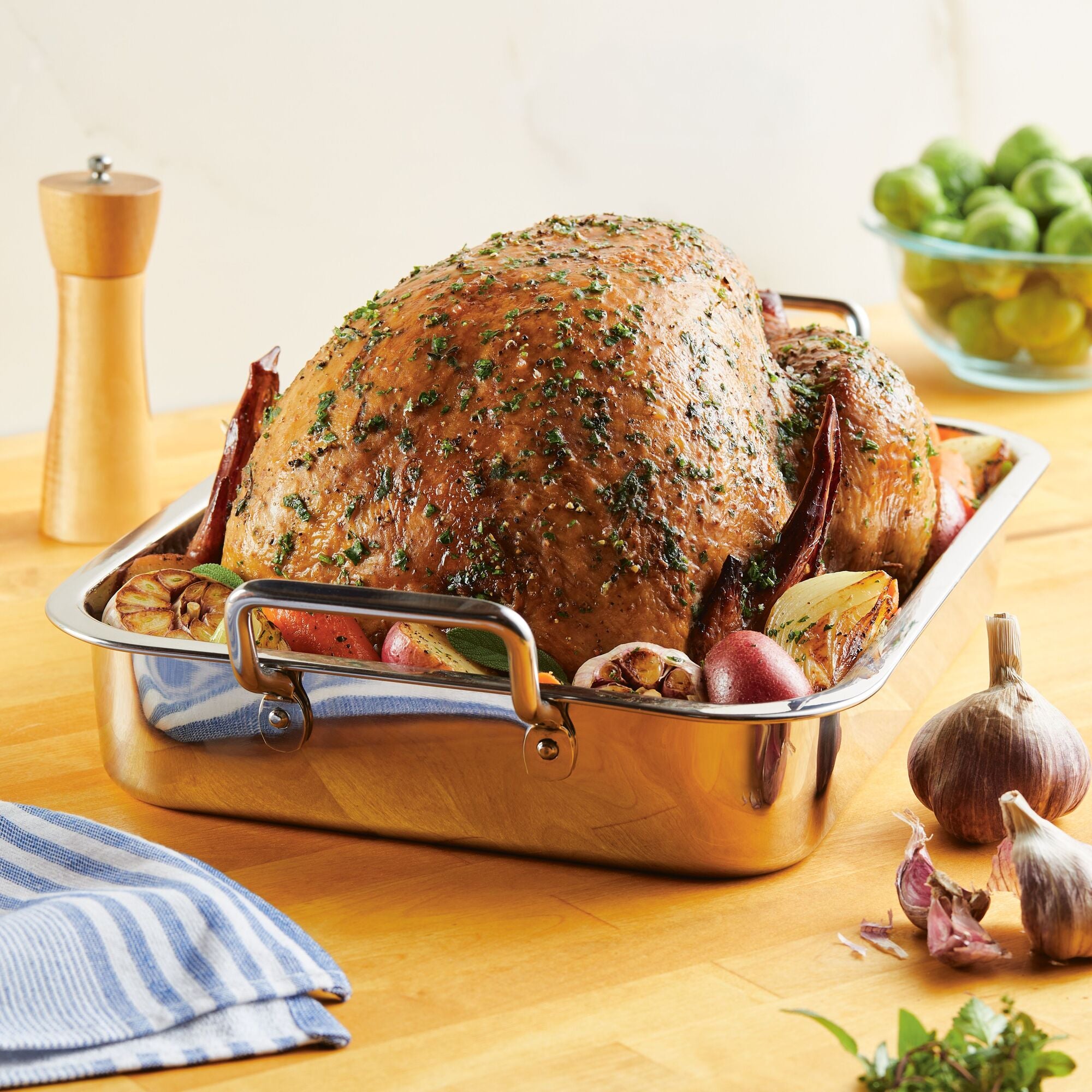 A roasted turkey sits in the 17" x 12.25" Stainless Steel Roaster with Rack, surrounded by garlic, onions, and herbs on a wooden table. This roaster features an oven-safe design and includes a removable rack for convenient serving.
