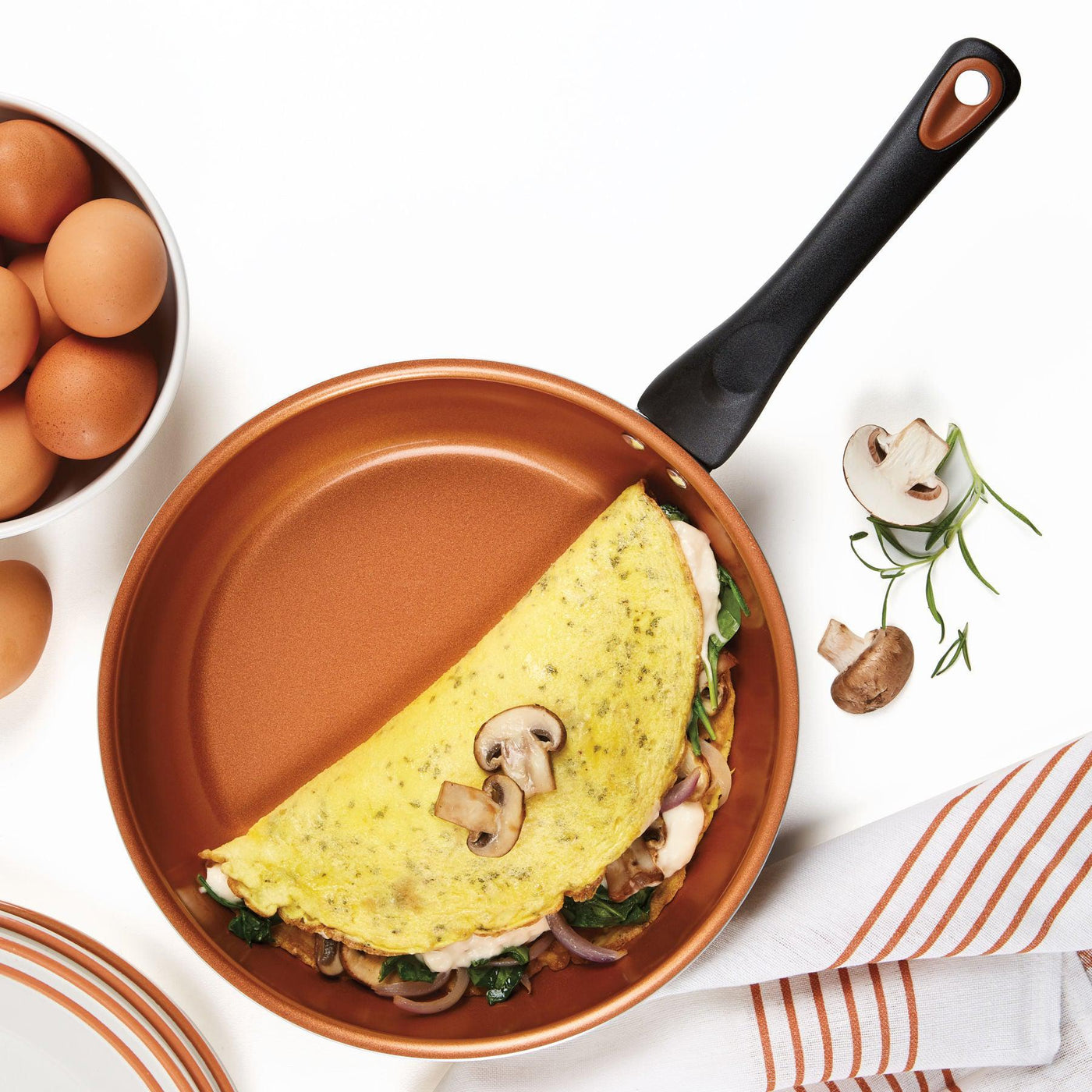 The omelette with mushrooms and spinach slides out effortlessly from the skillet, thanks to the innovative CopperSlide technology of the 12-Piece Copper Ceramic Nonstick Cookware Set, amidst a variety of kitchen items.