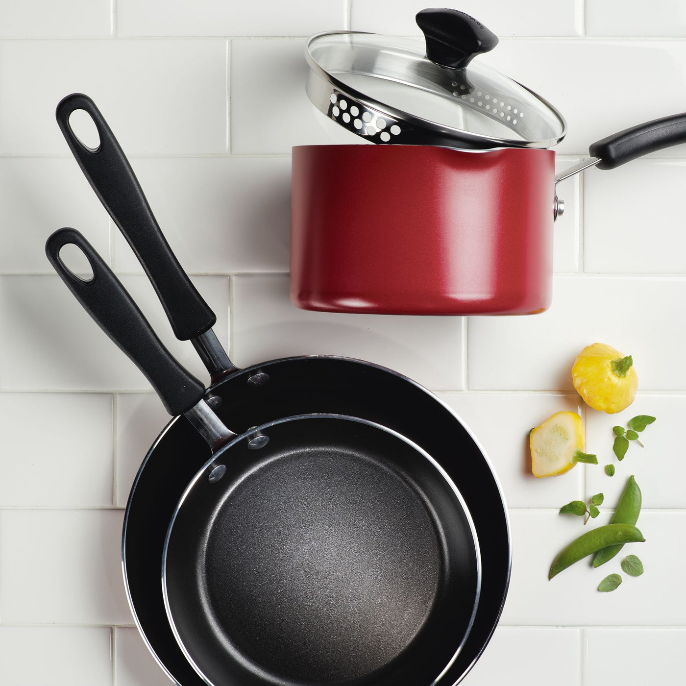 A red saucepan and two frying pans from the 15-Piece Nonstick Cookware Set, featuring DiamondMax technology, rest on a white tiled surface adorned with herbs.