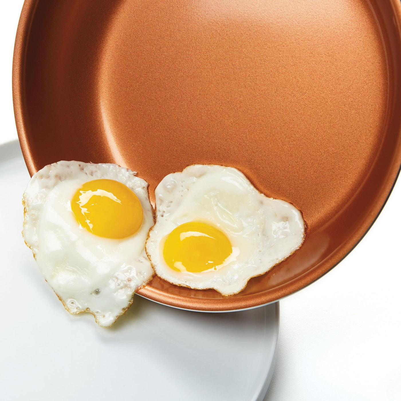Using CopperSlide technology, two fried eggs effortlessly slide off the 3-Quart Copper Ceramic Nonstick Straining Saucepan and onto a white plate.