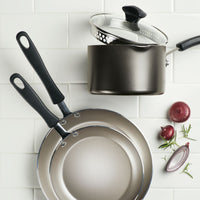 Part of the 15-Piece Nonstick Cookware Set, two frying pans crafted with heavy-gauge aluminum, a pot, onions, and herbs rest on a white tiled surface.