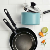 On a tiled surface, three pieces from the Nonstick Frying Pan Set sit alongside a lime, peppers, and sprigs of thyme. Among them is the standout frying pan, gleaming with its superior ceramic nonstick coating that guarantees effortless cooking and easy cleaning.