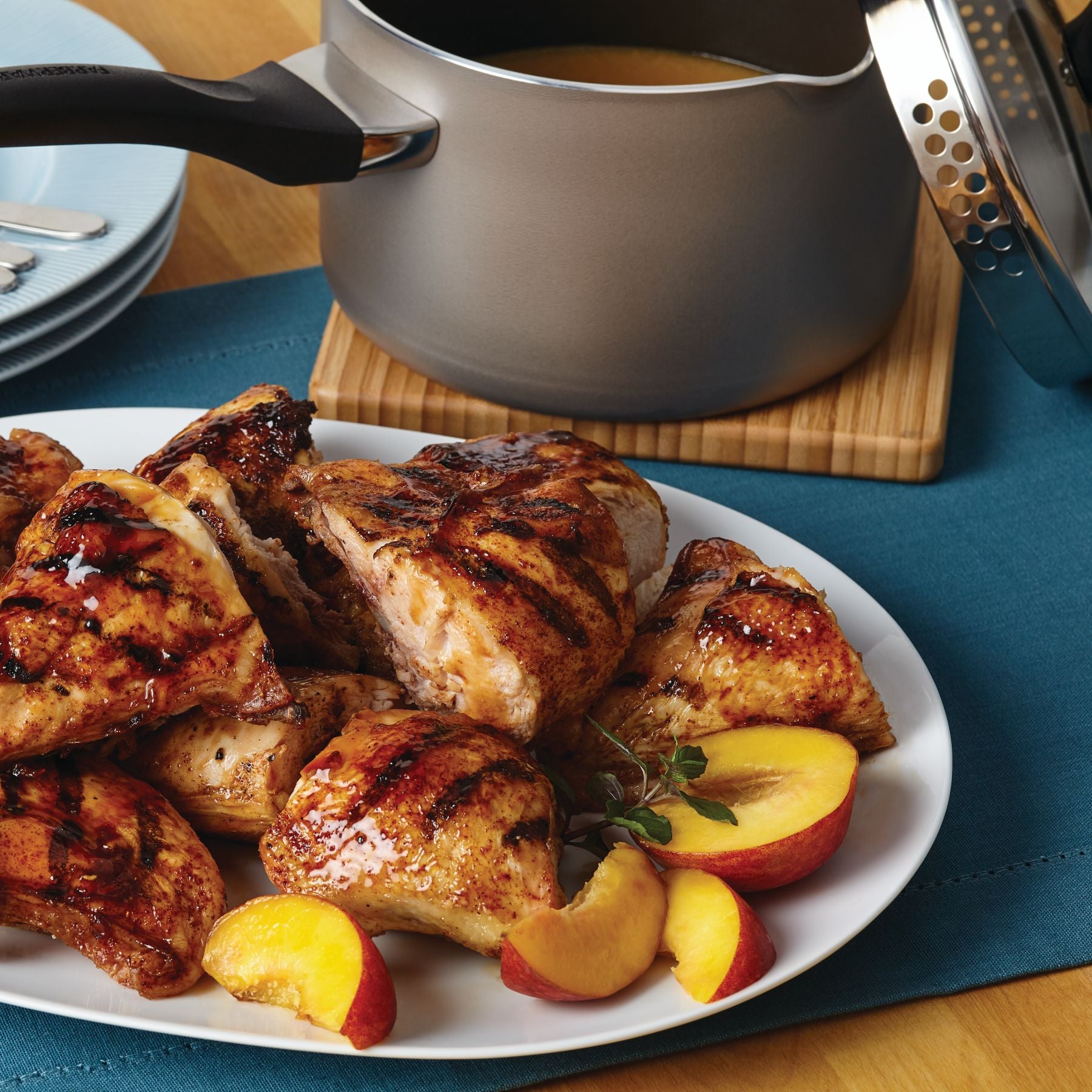 Grilled chicken with peach slices is elegantly presented on a plate next to the Nonstick Saucepan with Straining Lid, all set on a rustic wooden surface.