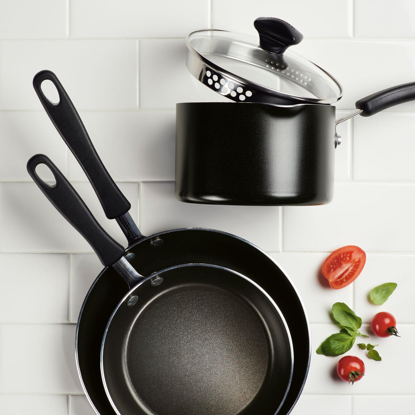 An 11-Inch Nonstick Square Griddle, two skillets, a saucepan with lid, tomatoes, basil, and garlic sit on white tiles.